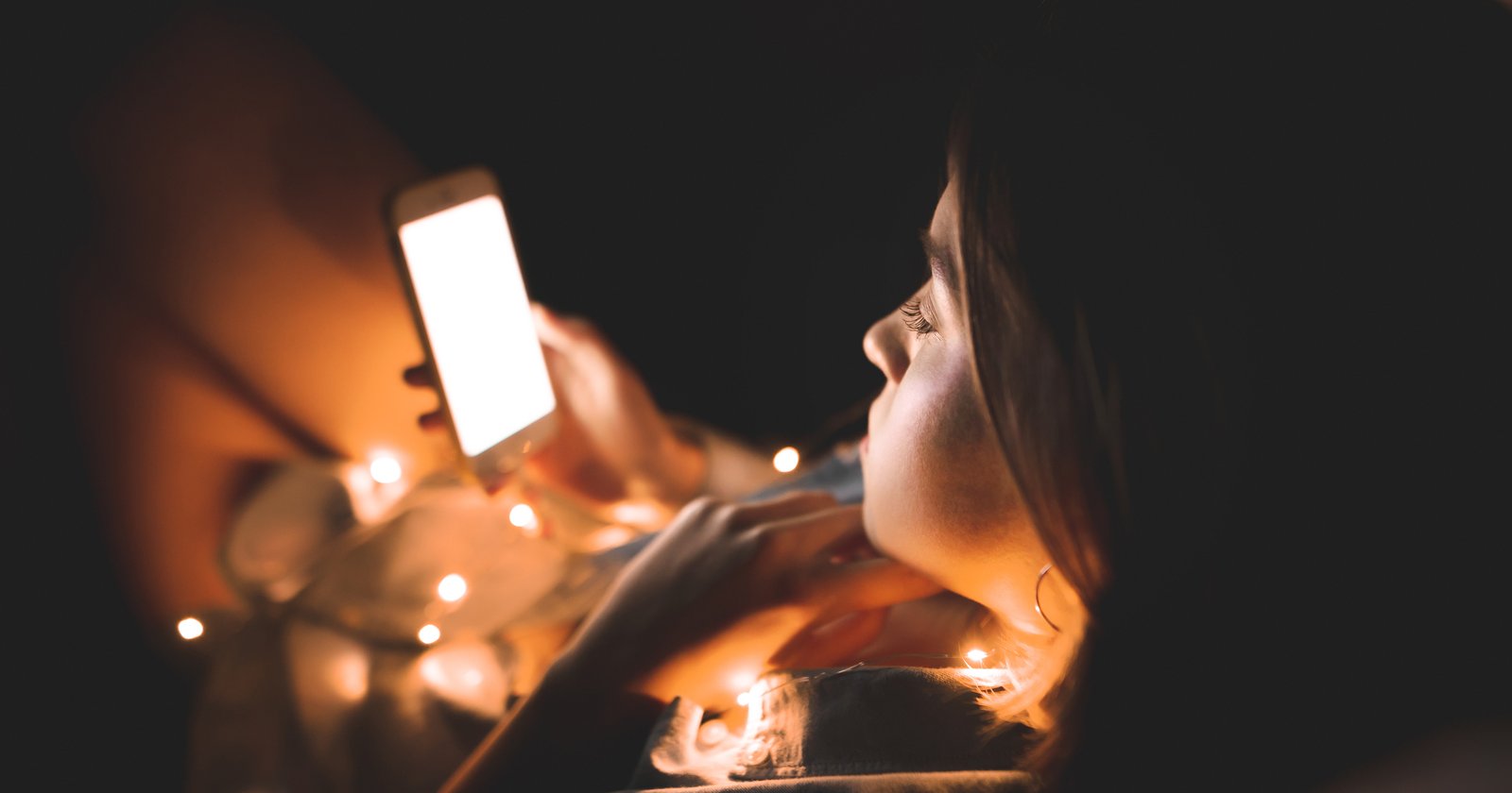Tânără întinsă pe pat, în întuneric, ținând un telefon aproape de față; ecranul aprins e singura sursă de lumină, iar în jurul ei sunt înșirate luminițe calde.